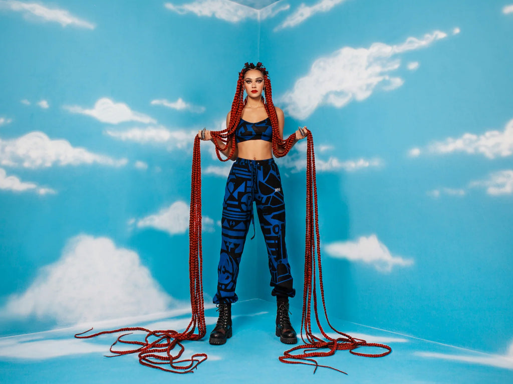 Model wearing BAM-BAM abstract cubist blue-and-black streetwear set with red braids, standing against a cloudy blue sky backdrop for the 'Blue is the New Black' campaign.